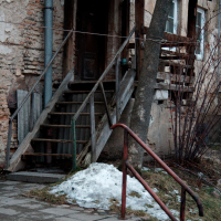 A weathered staircase leads to a dilapidated building entrance in Vilnius, with remnants of snow and bare branches hinting at the tail end of winter in an old urban environment.