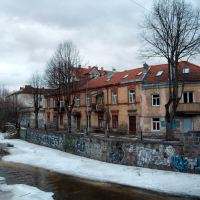 A row of weathered buildings with graffiti-covered bases in Vilnius overlooks a partially frozen river under a gloomy sky, encapsulating a serene yet urban decay ambiance.