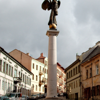 An angel statue perched on a tall column graces a quiet Vilnius street lined with buildings showing architectural pastels under a cloudy sky.