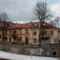 A row of aged, weathered buildings with graffiti-tagged walls stands beside a stone embankment in Vilnius, under a moody sky with bare trees signaling winter or early spring in a