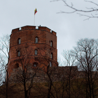A historic red brick tower stands atop a hill in Lithuania's capital, its battlements crowned with a fluttering national flag, while bare tree branches reach into the overcast sky, hinting at a