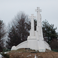 A trio of towering white crosses on a small, snow-dusted hill in Vilnius, Lithuania, flanked by bare trees under a cloudy sky, a symbol of faith and memorial standing solemn and