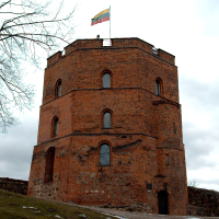 An imposing red-brick tower with a Lithuanian flag wafting at the top stands resolute against a cloudy sky in Vilnius, while patches of snow linger on the surrounding ground,