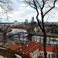 A cityscape of Vilnius viewed from a high vantage point, featuring a winding river, snow patches on the ground, leafless trees, and a mix of historic and modern buildings under a