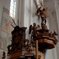 An ornate wooden pulpit adorned with religious sculptures and cherub figures, illuminated by the light filtering through a stained glass window in the background of a serene church interior in Vilnius.