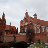 A historic church complex in Vilnius, showcasing gothic and baroque architecture, stands against a cloudy sky, with remnants of snow on the ground and a solitary statue in the foreground.