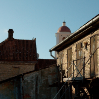 Old brick buildings in Vilnius with weathered facades under a clear blue sky, featuring exterior stairs and pipes, contrasted by the smooth dome of a distant building peering in the background.