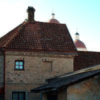 An old brick building with a red tiled roof stands in the foreground, while the domes of a historic structure in Vilnius, the capital city of Lithuania, peek out enigmatically in the