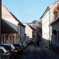 A quiet, narrow street in Vilnius, lined with parked cars and traditional buildings under a clear blue sky, with a solitary figure walking in the distance.