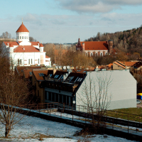 A European townscape under a clear blue sky, featuring historic architecture with a prominent white church tower, interspersed with more modern buildings and a sparse winter tree line in Vilnius.