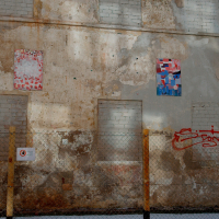 A weathered urban wall in Vilnius with patchy paint, featuring a chain-link fence in front, and adorned with assorted street art and graffiti including a red signature, a circular symbol, and