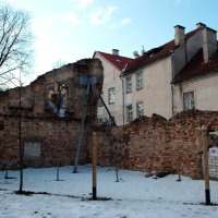The remains of a dilapidated brick wall, with the backdrop of intact buildings in Vilnius, suggest a contrast between destruction and preservation, possibly hinting at historical events or urban decay. Snow