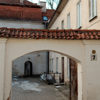 A weathered archway leads to a courtyard within an old European building complex in Vilnius, Lithuania, characterized by peeling paint, red roof tiles, and a rustic charm.