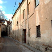 A narrow cobblestone alley winds between aged, weathered buildings with barred windows in Vilnius, evoking the historic charm of an old European town under a clear sky.