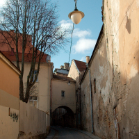 A lone street lamp dangles above a cobbled lane, flanked by old, weathered buildings with peeling façades, under a clear blue sky, in a quiet, historical European alley in