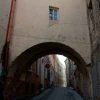 An archway spans over a narrow cobblestone street flanked by old buildings with textured facades under a clear blue sky in Vilnius, evoking a sense of history in a quaint urban