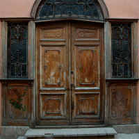 An ornate, weathered wooden double door with intricate ironwork, slight graffiti, and cobblestone steps set in an old building with a pink facade in Vilnius.