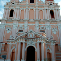 An imposing baroque-style church facade in Vilnius with ornate detailing, salmon-pink walls, two bell towers, and a grand staircase leading to the central entrance beneath a clear blue sky.