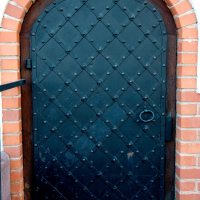 A sturdy, arched, black wooden door with a metal diamond pattern, sporting a ring knocker, set within a brick doorway in Vilnius, evoking a medieval or traditional European aesthetic.