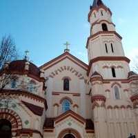 A towering church in Vilnius with a slender spire and reddish facade, featuring ornate arches and crosses, standing under a clear blue sky.