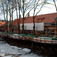 A winter's day view of a quaint Vilnius village with historic red-roofed houses, a pedestrian bridge under renovation, bare trees, and remnants of snow along the ground, hinting at