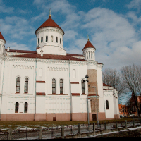 An imposing white cathedral with red-tiled roofs and multiple towers stands under a cloudy blue sky in Vilnius, partially surrounded by a wrought iron fence, showcasing classic architectural design.