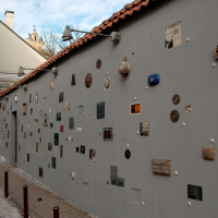 A quirky outdoor gallery in Vilnius with various small artworks, plaques, and sculptural elements affixed to a grey urban wall, slightly bathed in sunlight, alongside a cobbled street with