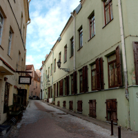 A narrow cobblestone street flanked by quaint, colorful buildings with weathered wooden shutters and doors, under a partly cloudy sky, in what appears to be a serene old town in Vilni