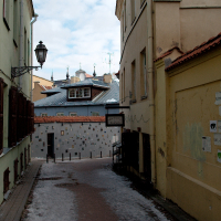 A snow-dusted alley in quaint Vilnius, characterized by narrow cobbled paths, ochre walls, antique street lamps, and the spire of a distant cathedral peering above tiled rooftops
