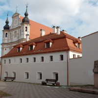 A baroque-style church towers behind a red-roofed building with white walls in Vilnius, complementing the quiet courtyard featuring benches and a bust monument under a serene blue sky with scattered clouds