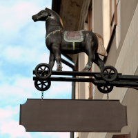 A vintage-style, three-dimensional horse figurine mounted on a wheeled platform, serving as a unique signboard against a building in Vilnius, Lithuania, with clear skies in the background.