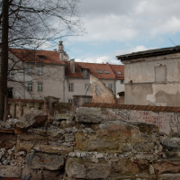 An old, dilapidated urban space in Vilnius with graffiti-covered walls, remnants of a crumbling brick fence in the foreground, and weathered buildings under a cloudy sky in the background, hint