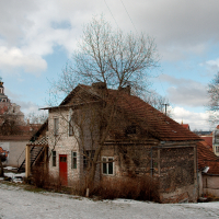 A dilapidated two-story building with graffiti art, a damaged roof, and overgrown vines stands in contrast to a cloudy sky and a distant cathedral's dome in Vilnius, highlighting urban decay