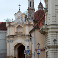 A Baroque-style gate with elaborate sculptural details leads to a cobblestone street flanked by classic European architecture under a cloudy sky in Vilnius, evoking a historic ambiance.