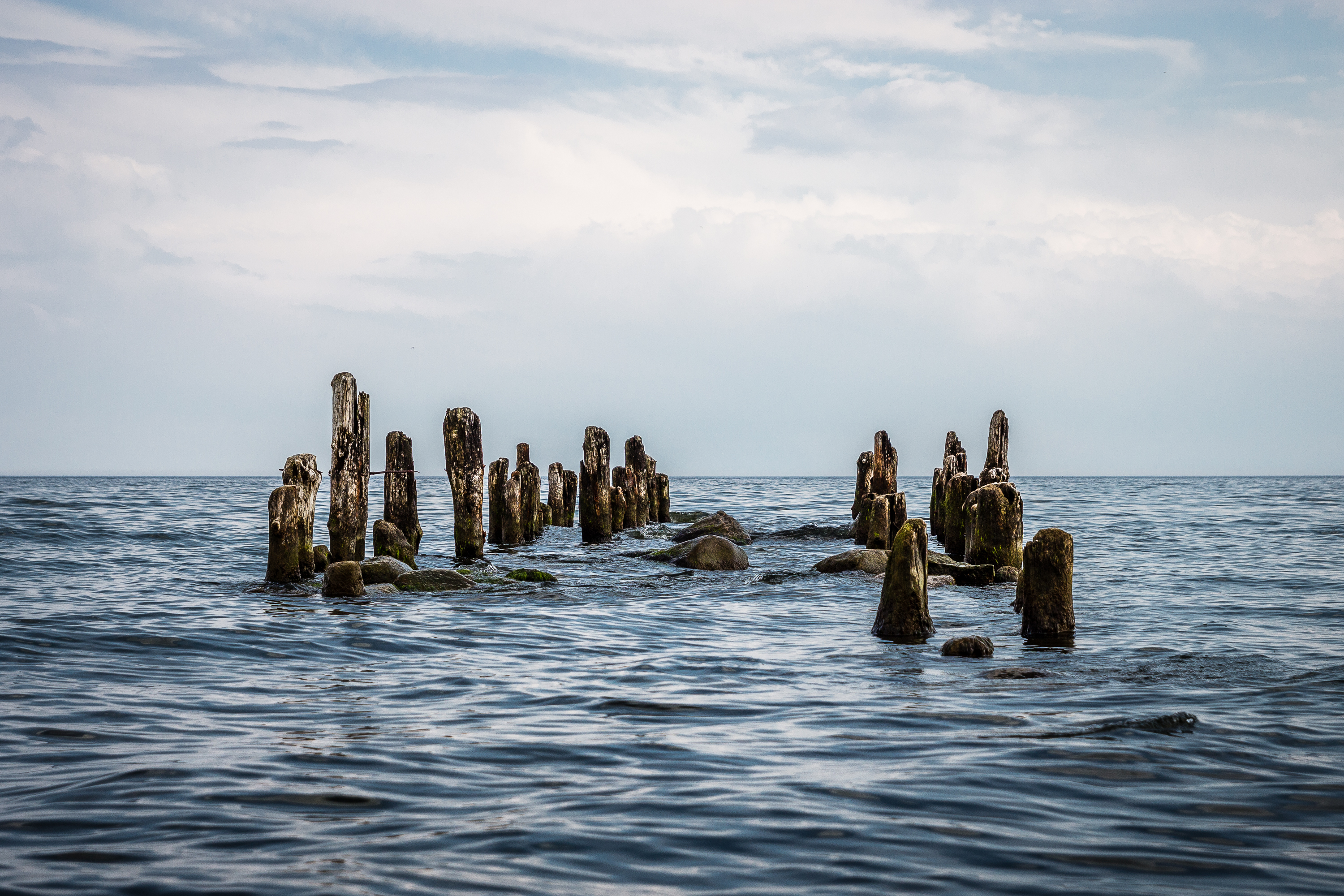 A serene seascape with partially submerged weathered wooden posts forming the remnants of an old pier, emerging from calm, dark blue waters under a cloudy sky. Scattered rocks are visible among the posts, and the horizon blends seamlessly into the sky. via @atisgailis