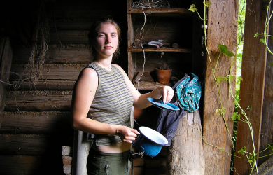 A Woman Holding A Blue Bowl In Front Of A Wooden Hut During Summer At A Country House.